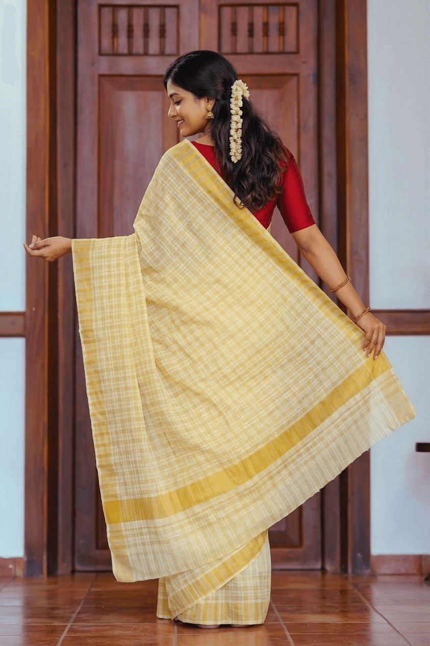 A woman wearing a light yellow and white checkered saree with a red blouse, standing in front of a dark wooden door and holding out the saree's pallu to display its pattern and texture.