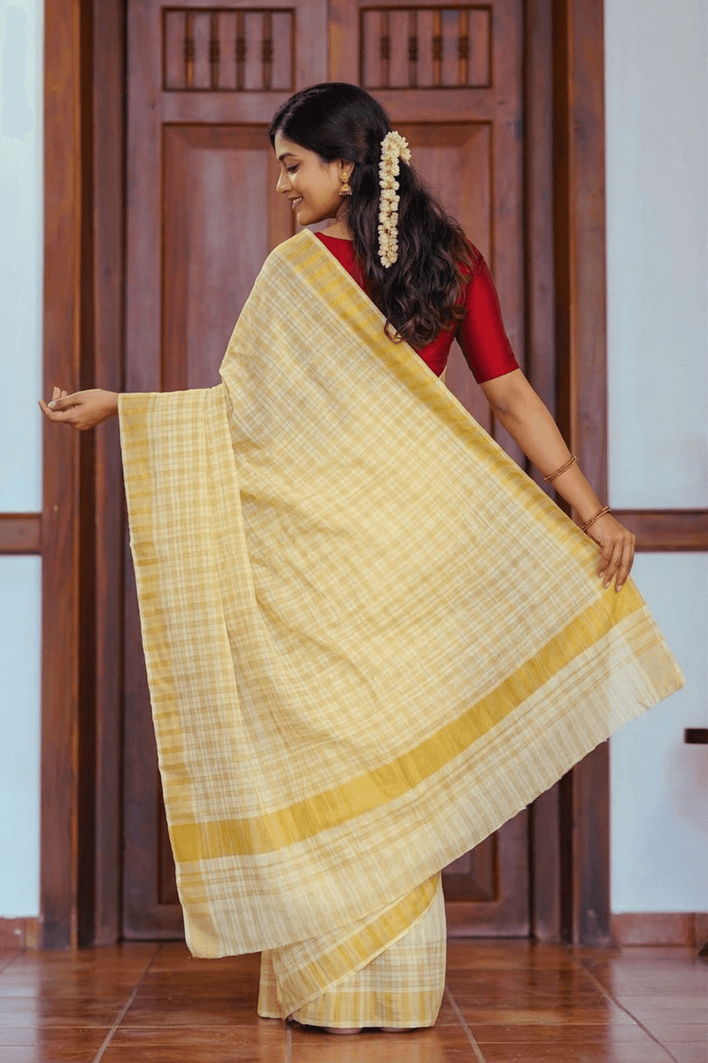 A woman wearing a light yellow and white checkered saree with a red blouse, standing in front of a dark wooden door and holding out the saree's pallu to display its pattern and texture.