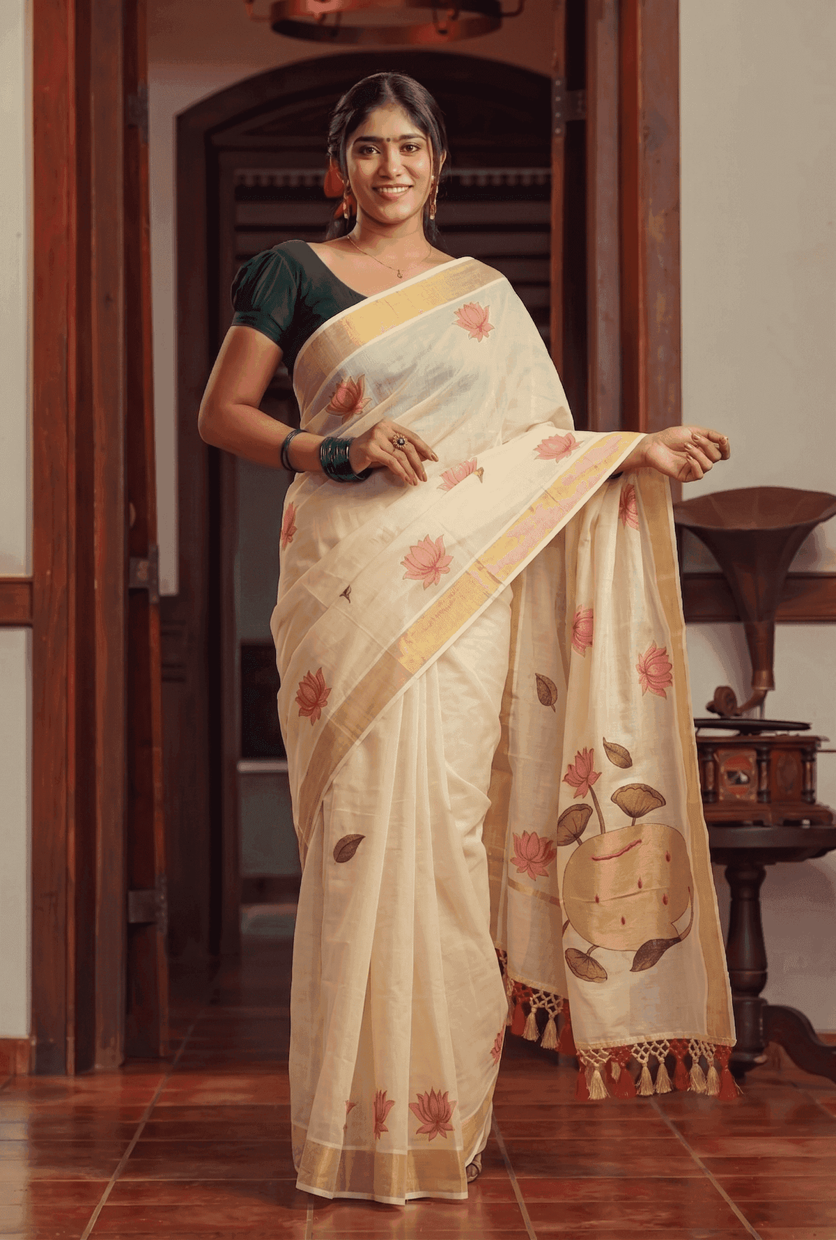 A woman wearing a cream-colored cotton saree with pink floral prints, a gold border, and a dark green blouse, standing in a traditional indoor setting with wooden doors and tiled floors.