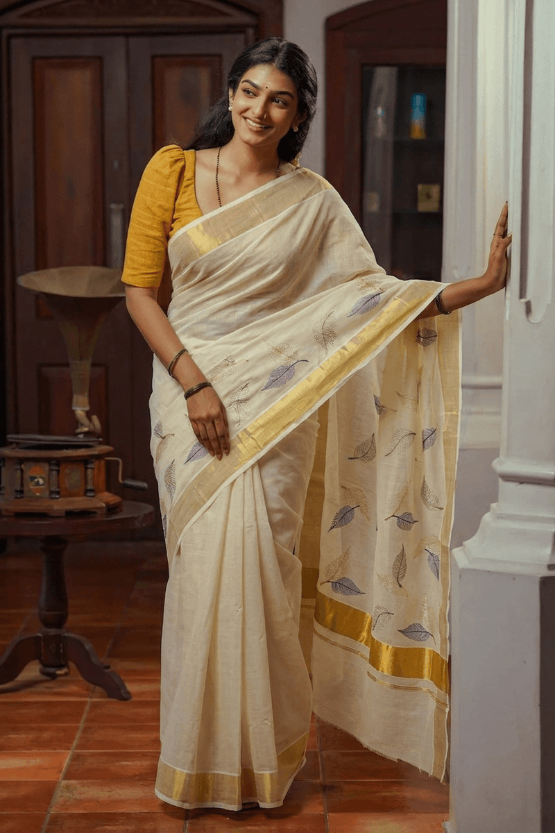 A woman wearing a cream-colored silk saree with a yellow blouse, standing in a traditional room with wooden furniture and tiled floors.