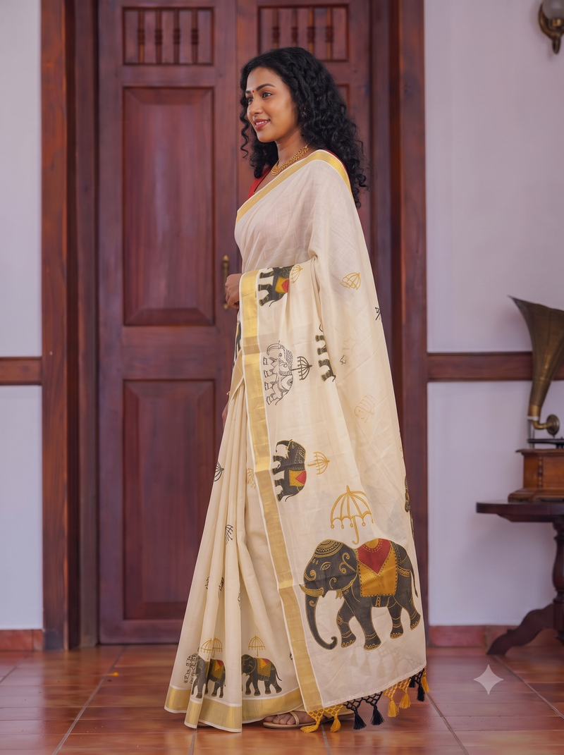 A woman wearing a cream-colored saree with elephant and traditional motif prints, standing in front of a dark wooden door in an indoor setting.