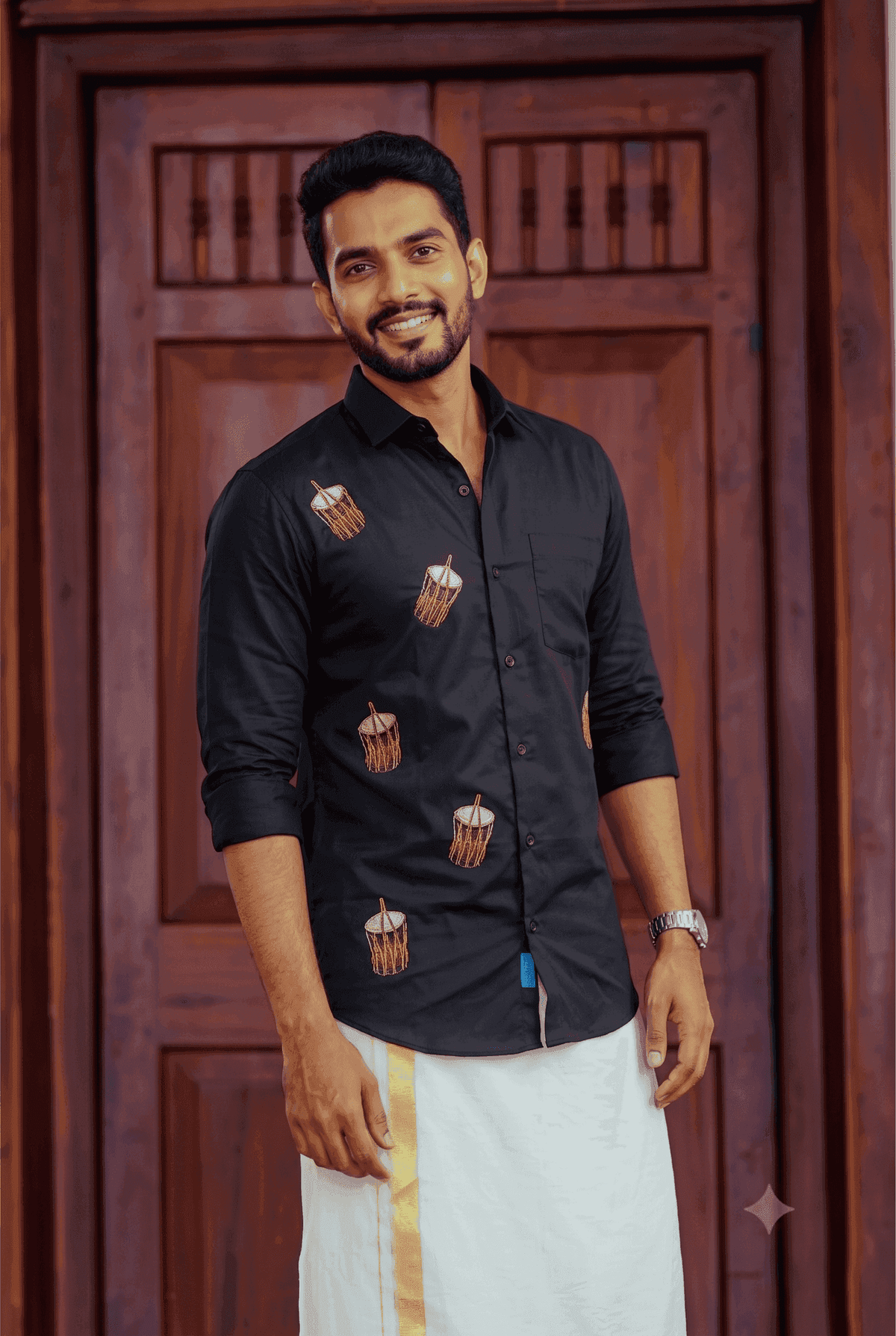 A man wearing a black long-sleeve button-up shirt with small brown and white drum illustrations, paired with a white traditional garment, standing in front of a dark wooden door.