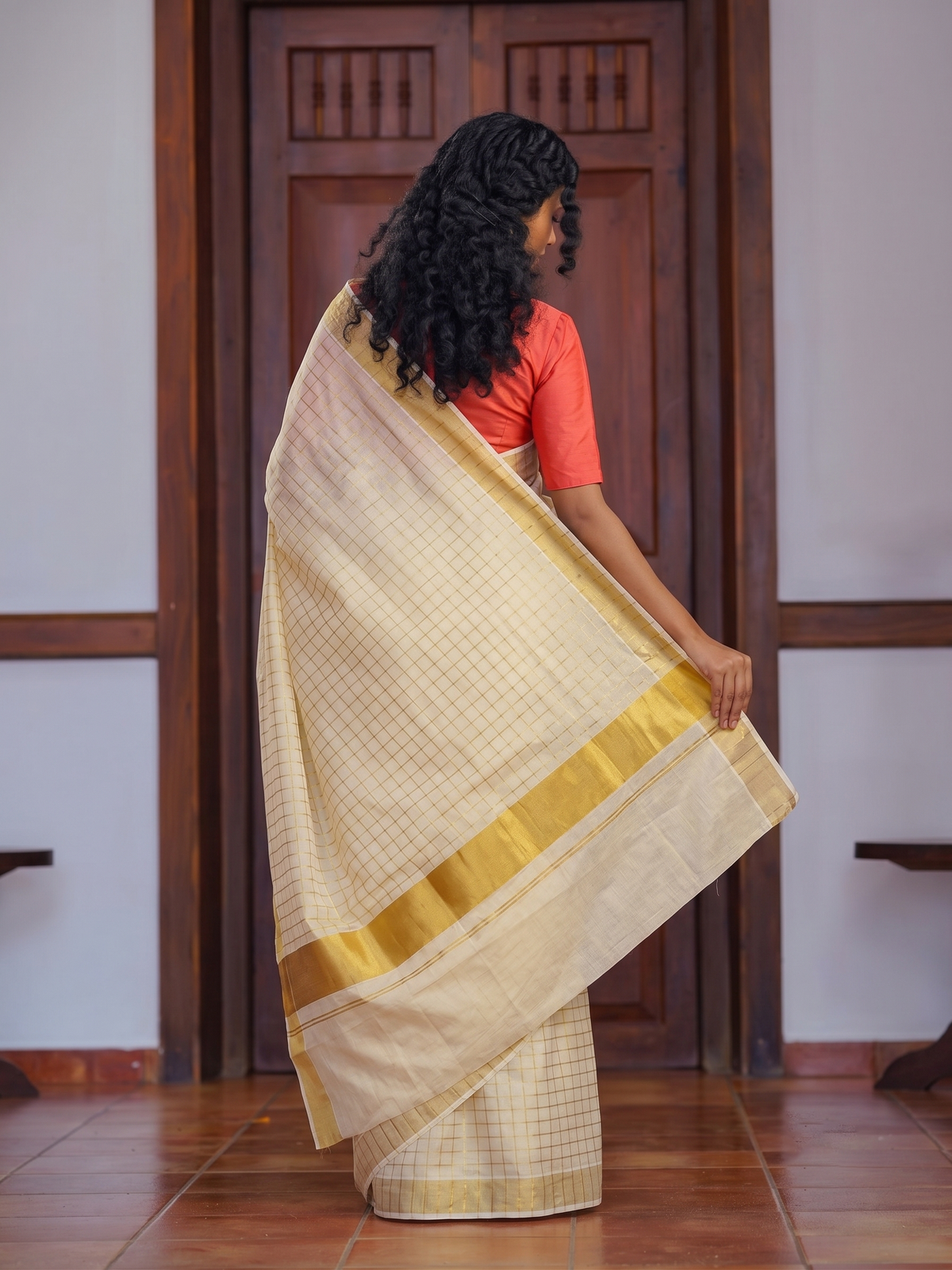 A woman wearing a cream-colored checkered saree with a gold border and a coral orange blouse, standing in front of a dark wooden door.