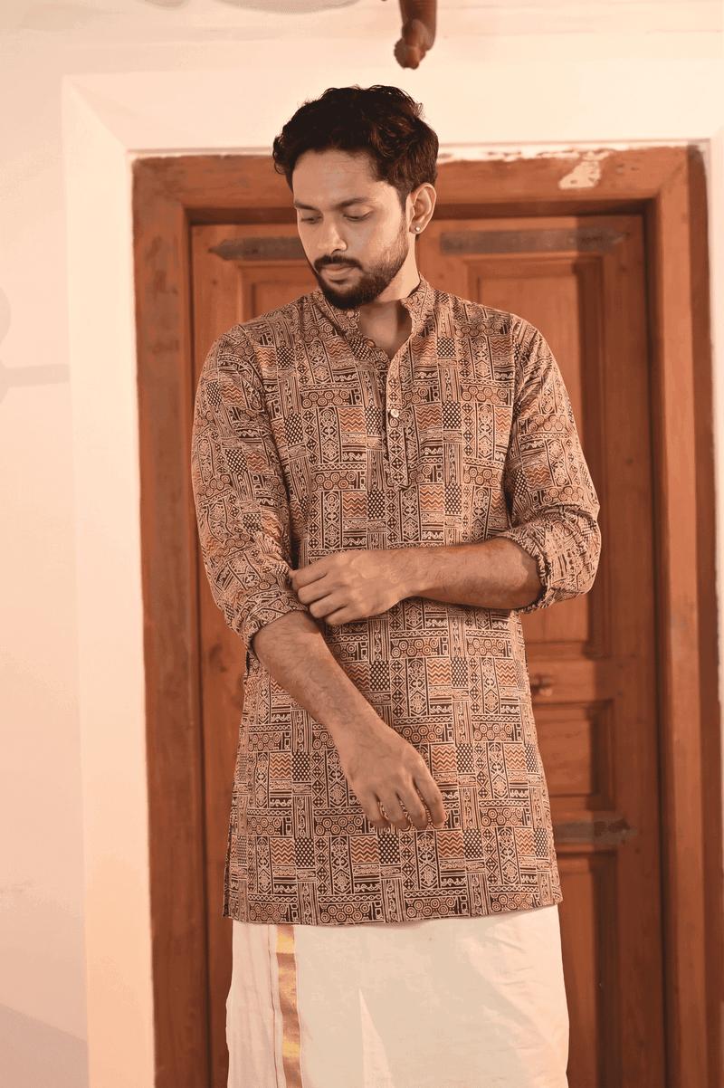 A man wearing a brown and beige patterned kurta with a white dhoti, standing in front of a wooden door.
