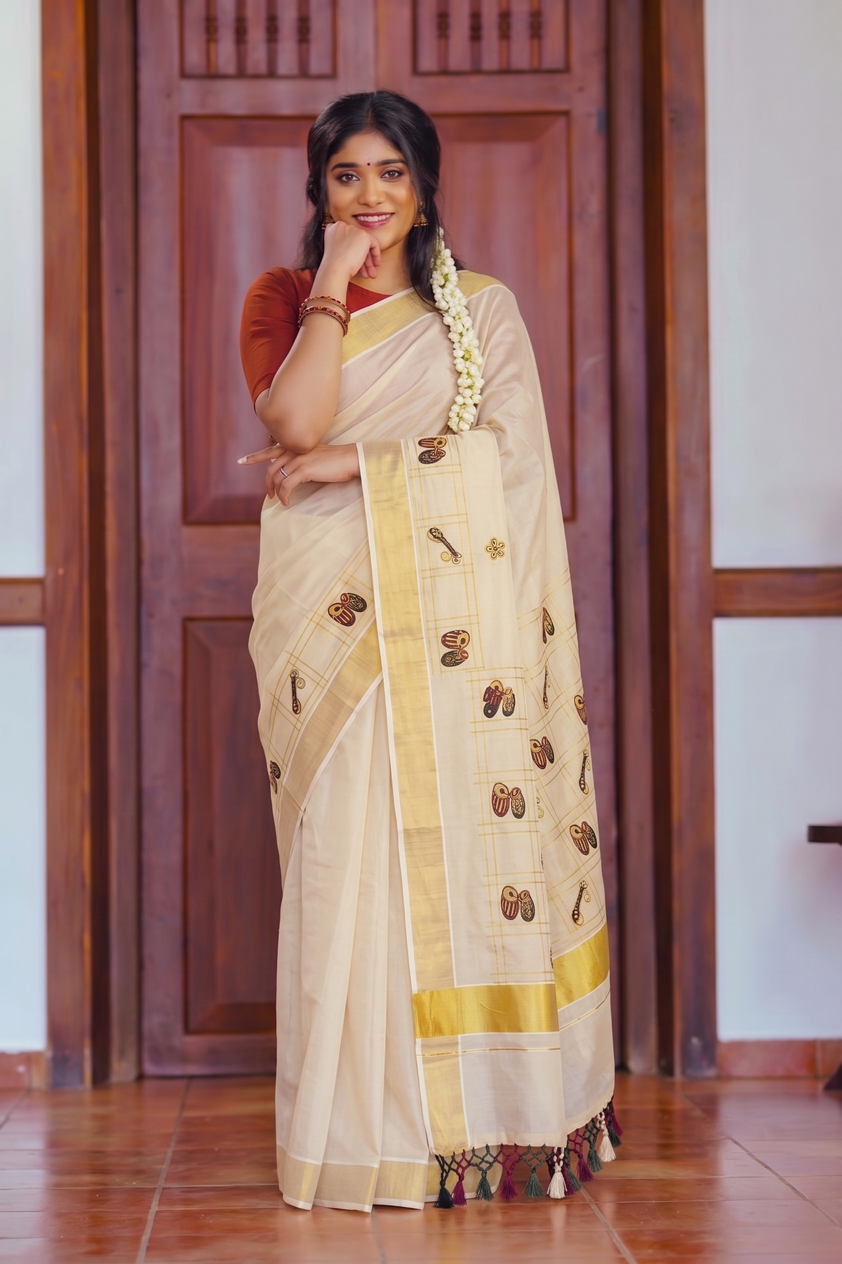 A woman wearing a traditional Indian saree stands in front of a wooden door. The saree is cream-colored with a gold border and small brown floral motifs, paired with a rust-red sleeve.
