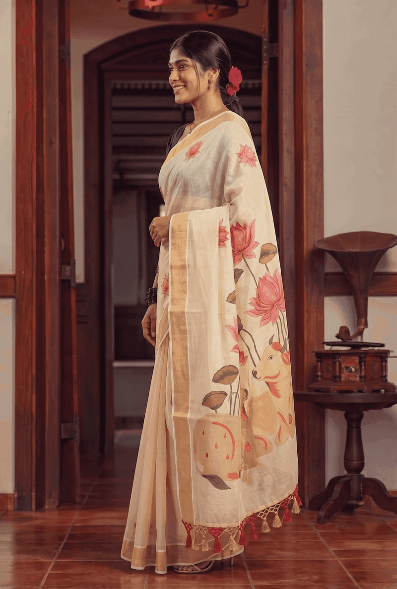 A woman wearing a cream-colored saree with pink lotus floral prints and green leaves, standing in a traditional indoor setting with wooden doorframes and a vintage gramophone.