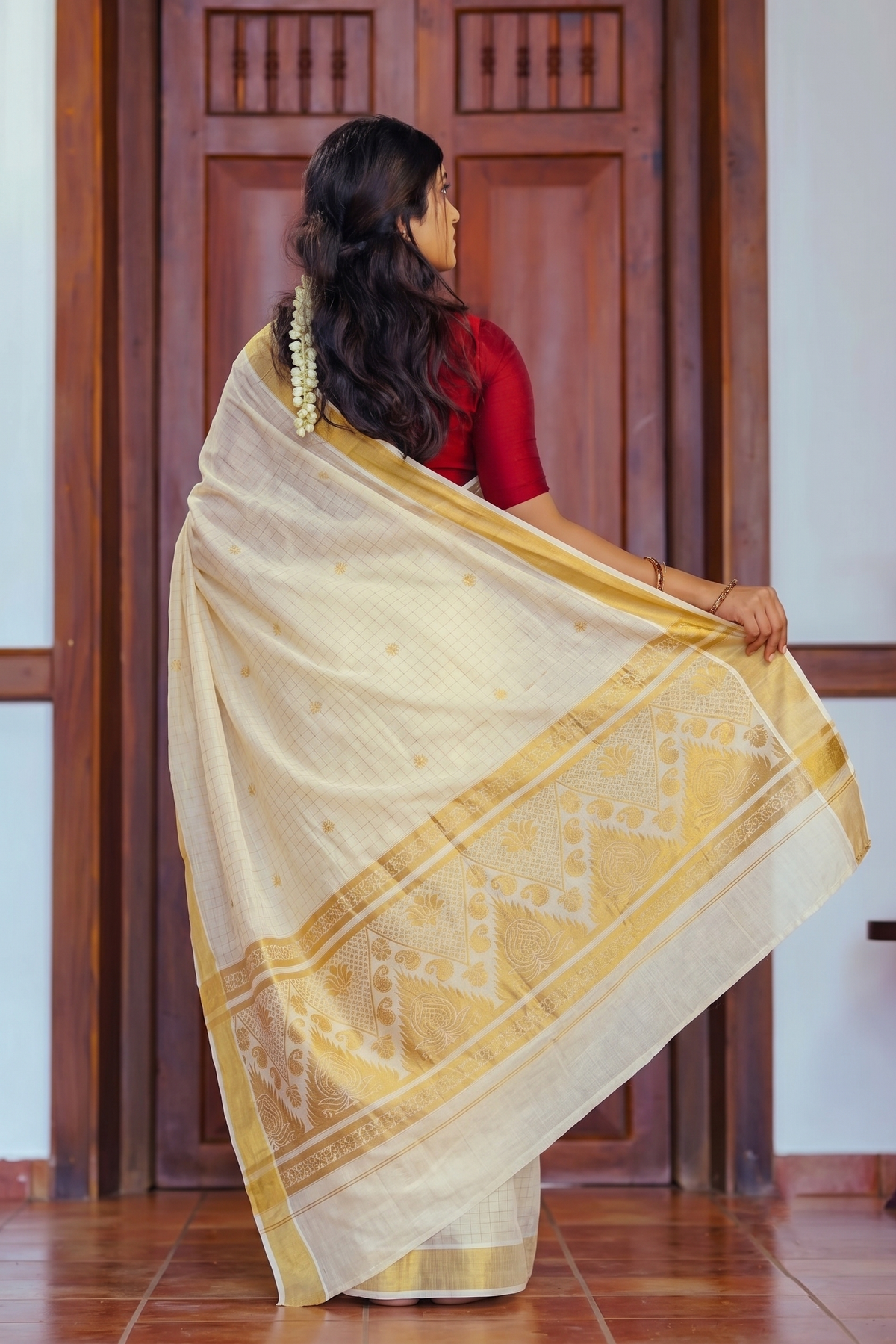 A woman wearing a cream-colored silk saree with a yellow border and a red blouse, standing in front of a wooden door and holding out the saree drape.