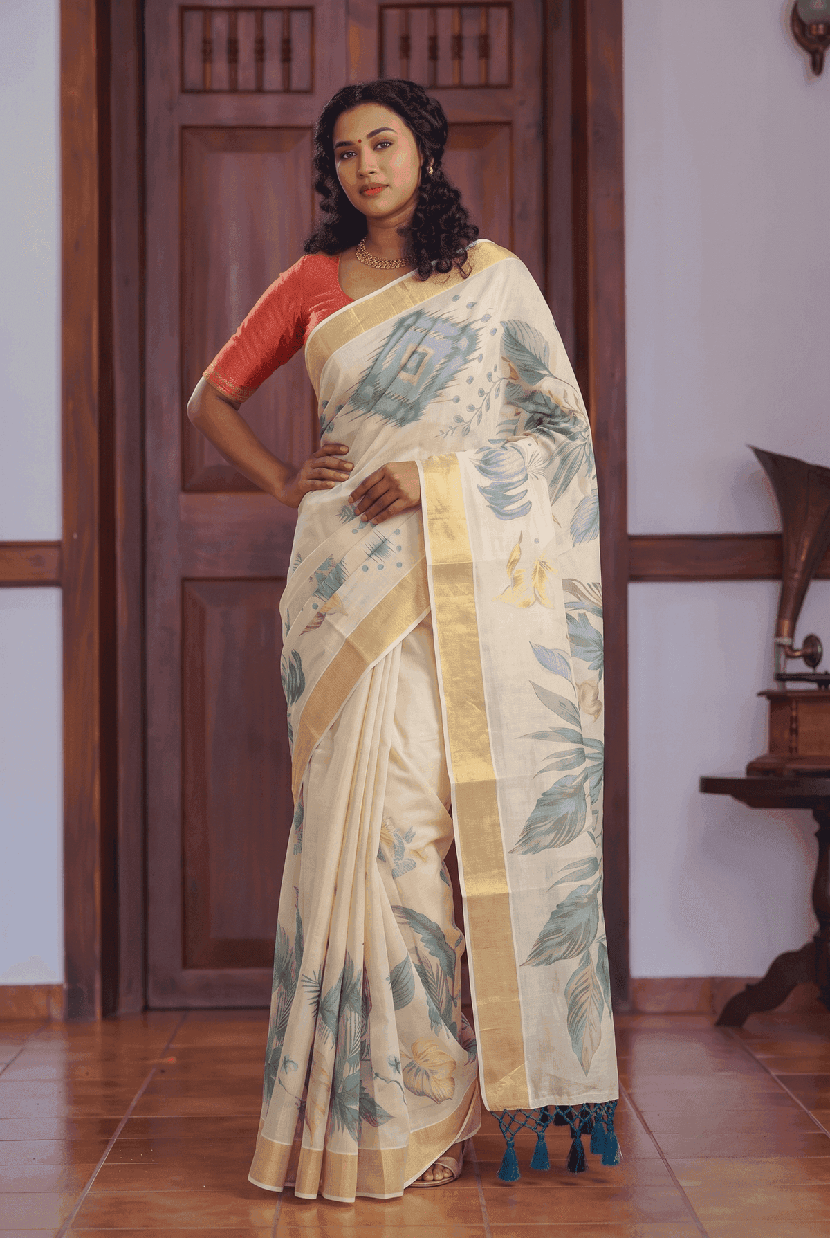 A woman models a cream-colored printed saree with blue-green leaf motifs and a gold border, paired with a red-orange sleeveless blouse, standing in front of a wooden door.
