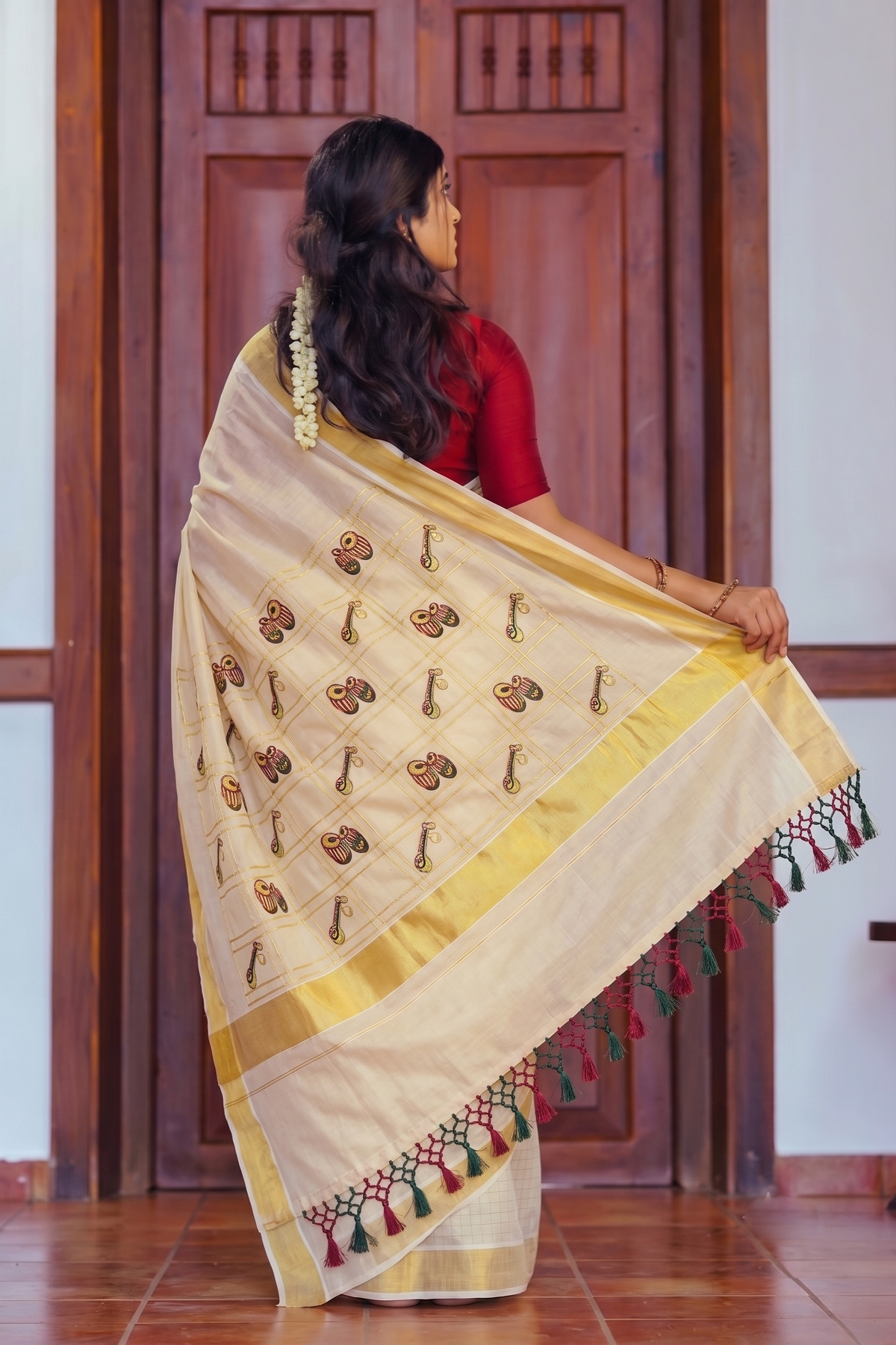 A woman wearing a cream-colored silk saree with gold borders and brown floral motifs, paired with a red blouse, standing in front of a wooden door.