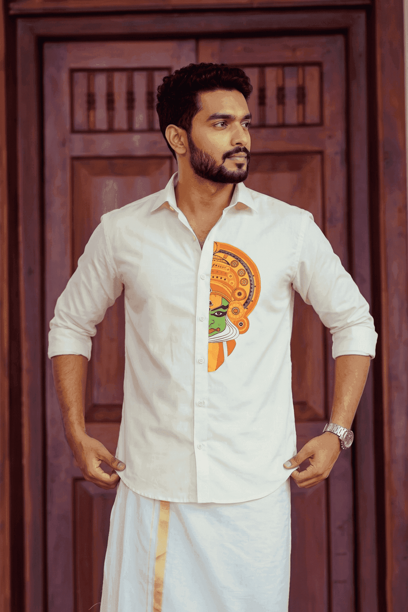 A man wearing a cream-colored cotton kurta with a colorful traditional Indian motif on the front, paired with a white dhoti, standing in front of a dark wooden door.