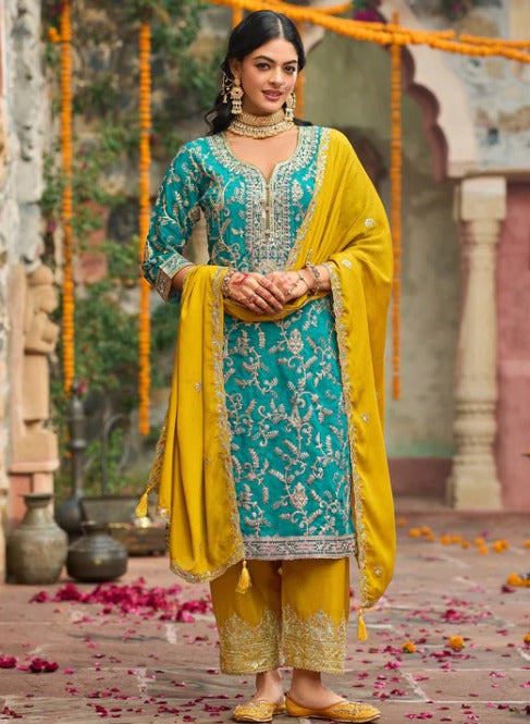 A woman models a traditional Indian salwar kameez suit set featuring a turquoise blue embroidered kurta with gold zari work, matching yellow trousers, and a yellow dupatta, set against an ornate courtyard background.