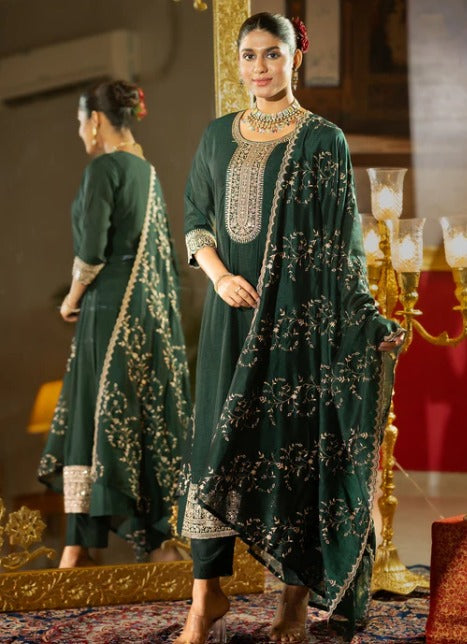 A woman models a dark green traditional outfit with intricate gold embroidery, shown both from the front and reflected in a mirror. The setting features warm, golden lighting and traditional decor.