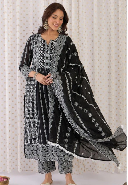 A woman modeling a black three-piece ethnic suit with intricate white embroidery and a matching dupatta, set against a light patterned curtain backdrop.
