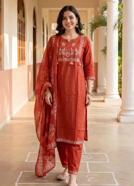A woman stands in a sunlit corridor wearing a rust-orange traditional outfit with matching pants and a sheer printed dupatta. The outfit has white embroidered detailing on the chest and sleeve cuffs.