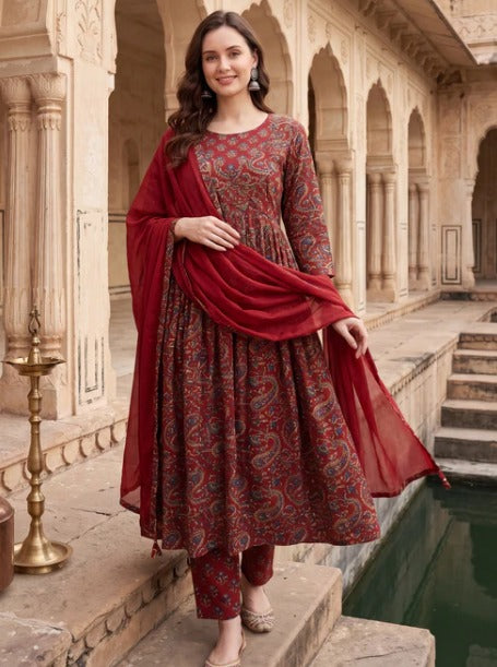 A woman models a traditional Indian salwar kameez set in a courtyard with stone arches and a pool. The outfit features a deep red base with intricate paisley patterns in blue, gold, and green, complete with a matching sheer red dupatta.