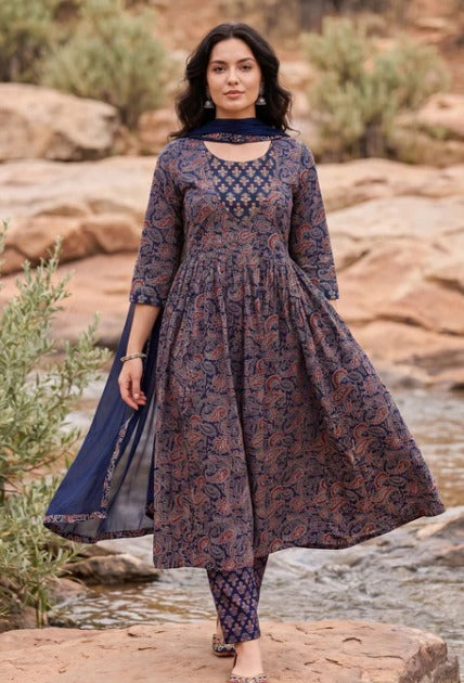 A woman wearing a dark navy blue and brown paisley printed kurta set with matching leggings and a dupatta, standing outdoors on a rocky stream bed.