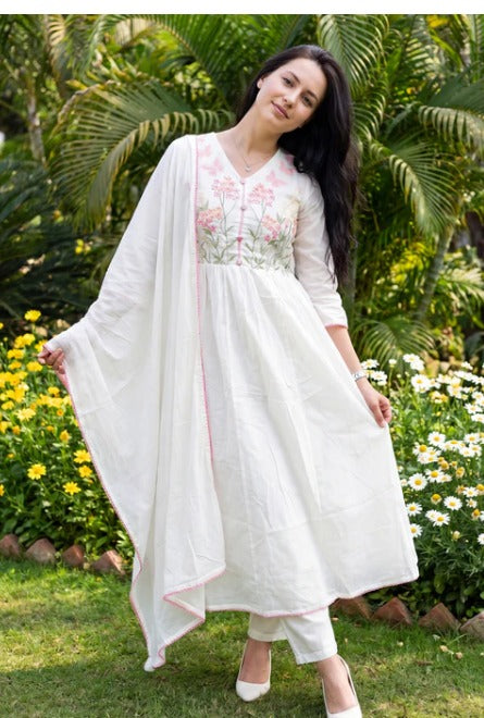 A woman wearing a white embroidered kurta set with a matching sheer drape, standing outdoors in a garden with green foliage and flowers.