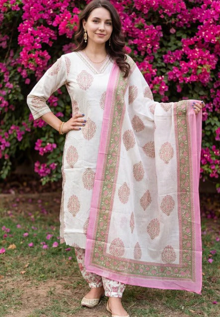A woman modeling a traditional Indian salwar kameez suit set in an outdoor garden setting with pink bougainvillea flowers. The set features an off-white kurta and matching trousers with pink and green printed motifs, paired with a pink border dupatta.
