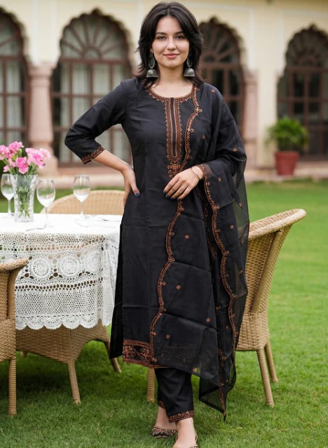 A woman stands outdoors on a lawn wearing a black three-piece traditional outfit with brown embroidered details. The set includes a long kurta, matching pants, and a dupatta draped over her arm.