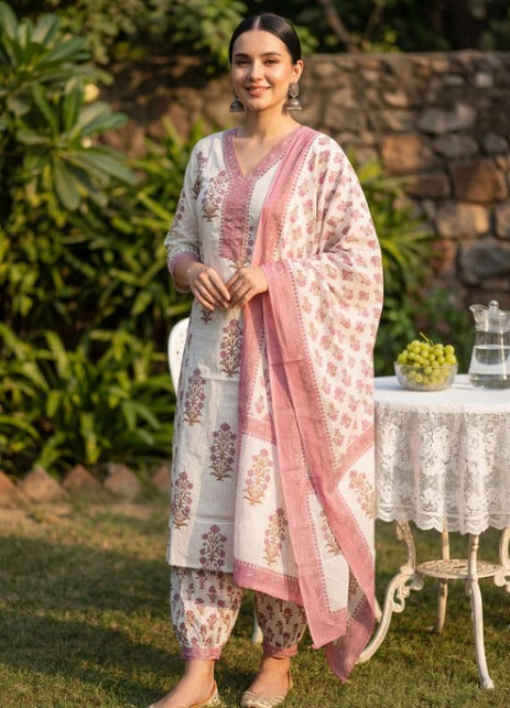 A woman stands outdoors in a garden setting wearing a traditional three-piece salwar kameez suit. The suit features an off-white kurta and matching trousers with dusty pink floral block prints, paired with a solid pink dupatta.