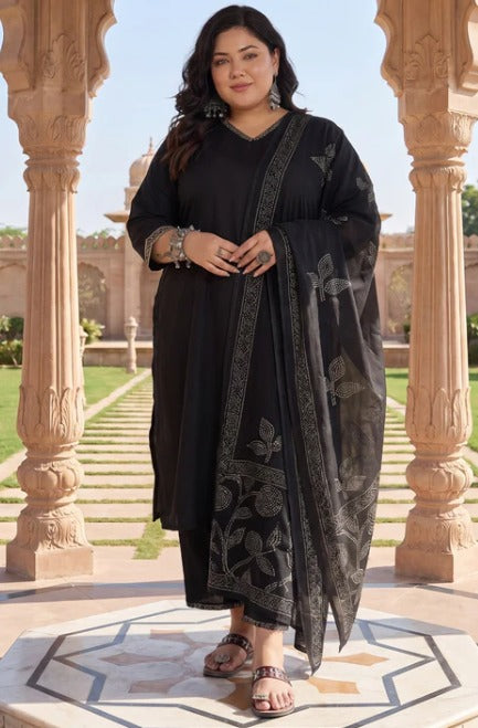 A woman modeling a black kurta set with a matching printed dupatta, standing between ornate stone pillars in an outdoor courtyard.
