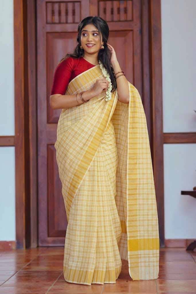 A woman wearing a yellow and white checkered saree with a red blouse, standing in front of a wooden door.
