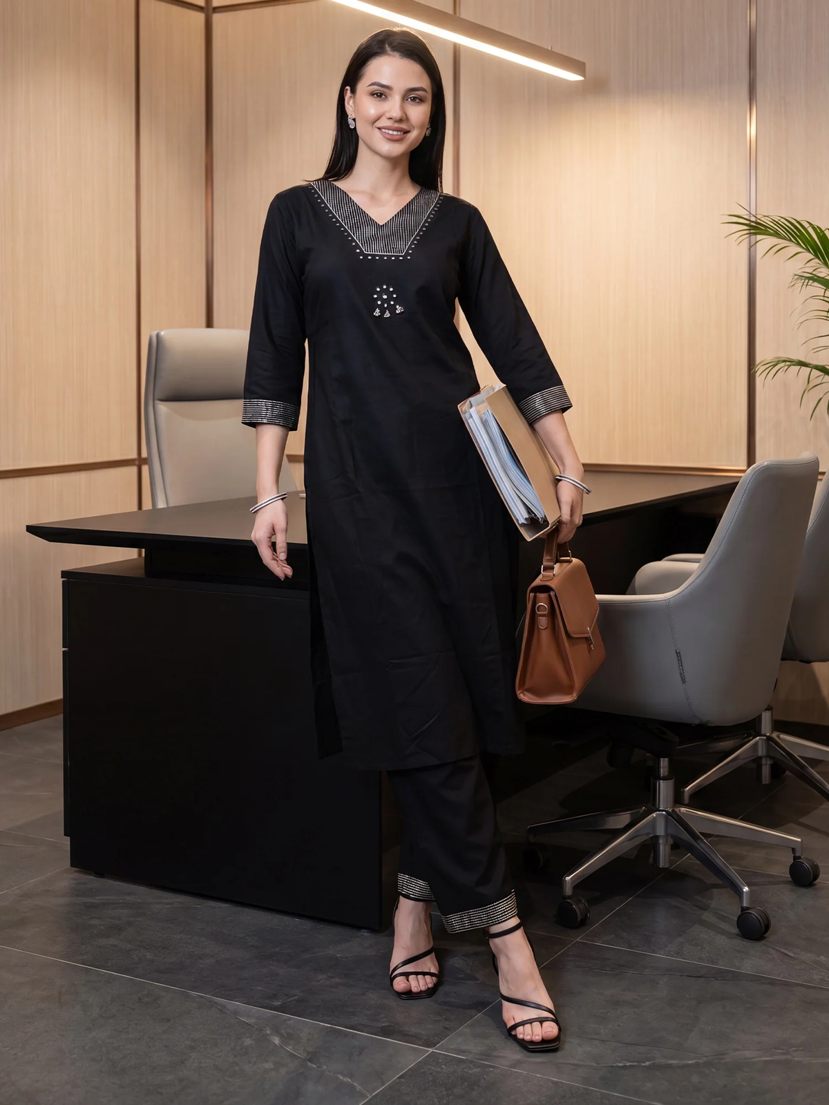 A woman models a black three-piece kurta set with grey and silver accents in a modern office setting. The set includes a long-sleeved kurta with a V-neckline, matching trousers, and grey cuffs.