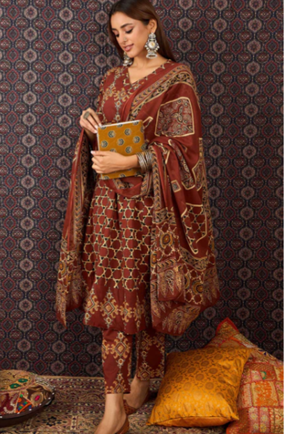 A woman models a traditional South Asian salwar kameez suit in maroon with intricate gold and beige geometric patterns, paired with a matching dupatta and holding a small gold clutch, set against a dark patterned wall.