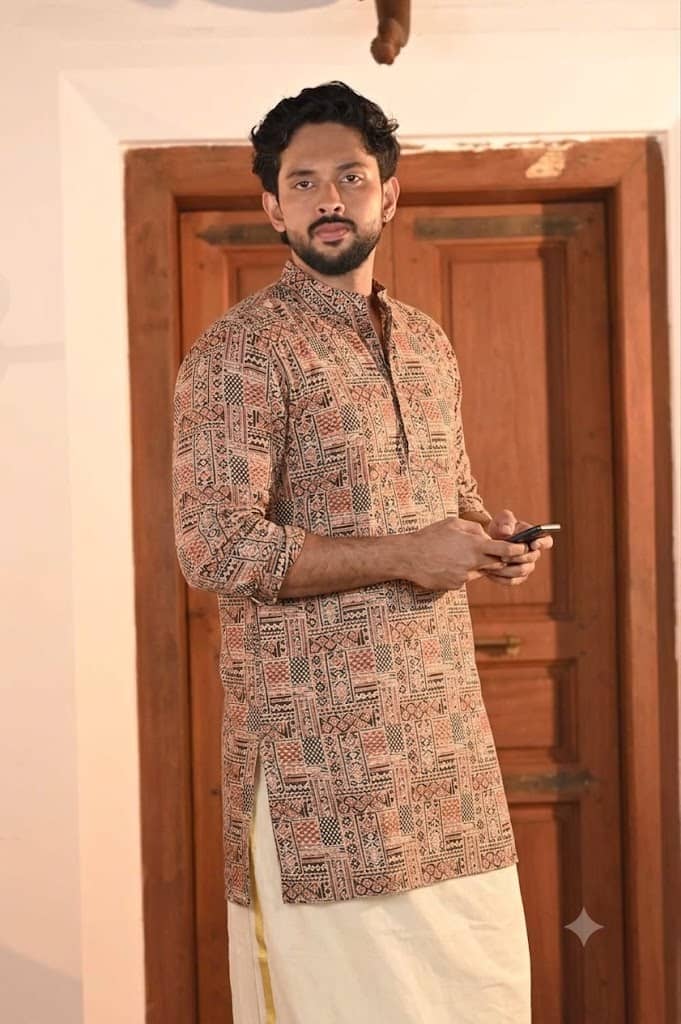 A man wearing a patterned beige and brown kurta with a white dhoti, standing in front of a wooden door.