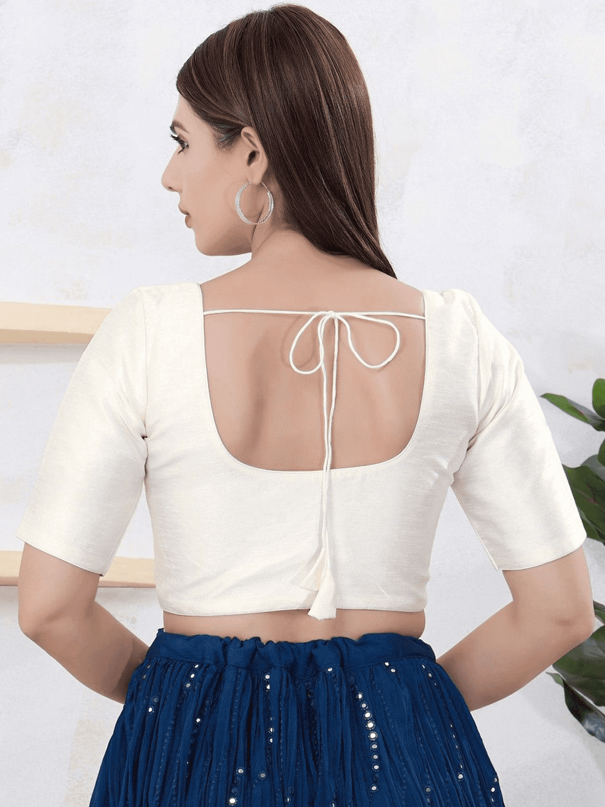 A woman shown from the back wearing a cream-colored short-sleeve blouse with a tie-back neckline and a dark blue pleated skirt with small silver embellishments.