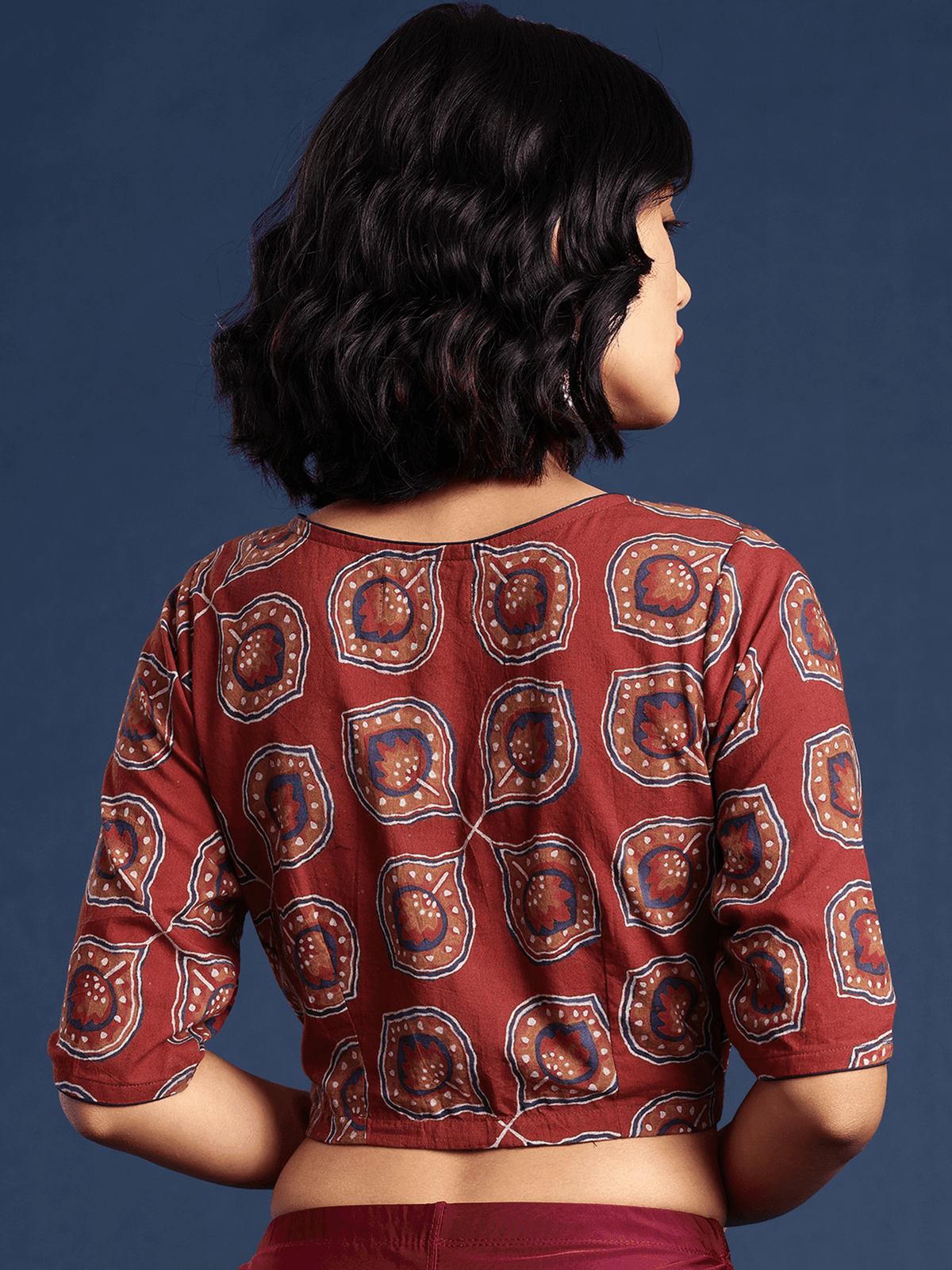 A woman shown from the back wearing a rust-red cropped blouse with a repeating paisley-style print in brown, gold, and blue, paired with matching red bottoms, set against a dark blue studio background.