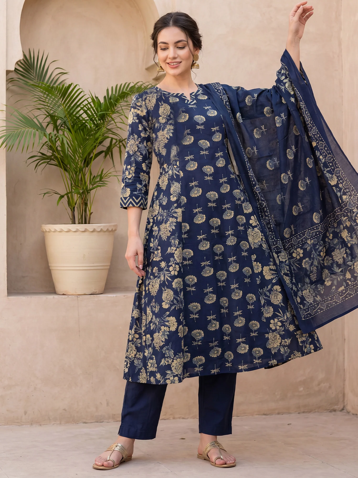 Woman wearing a blue floral outfit with a matching dupatta, standing against a beige wall with a plant.