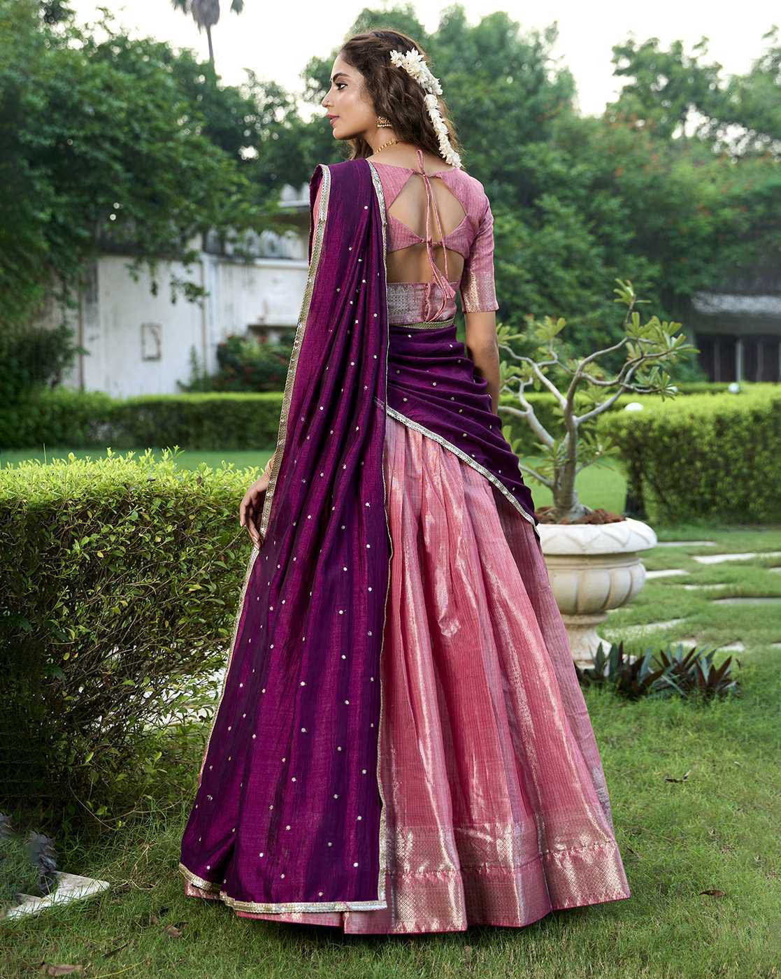 Back view of model wearing women's woven lehenga choli set in pink silk skirt and purple dupatta with tie back blouse