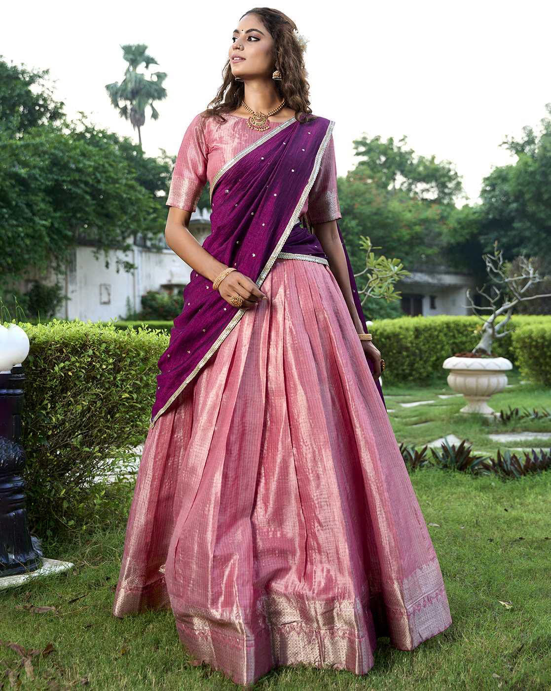Model wearing a pink silk women's woven lehenga choli set with purple dupatta and gold border in garden