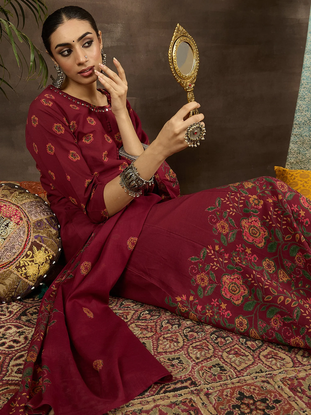 Woman in a maroon saree with floral patterns holding a decorative mirror.