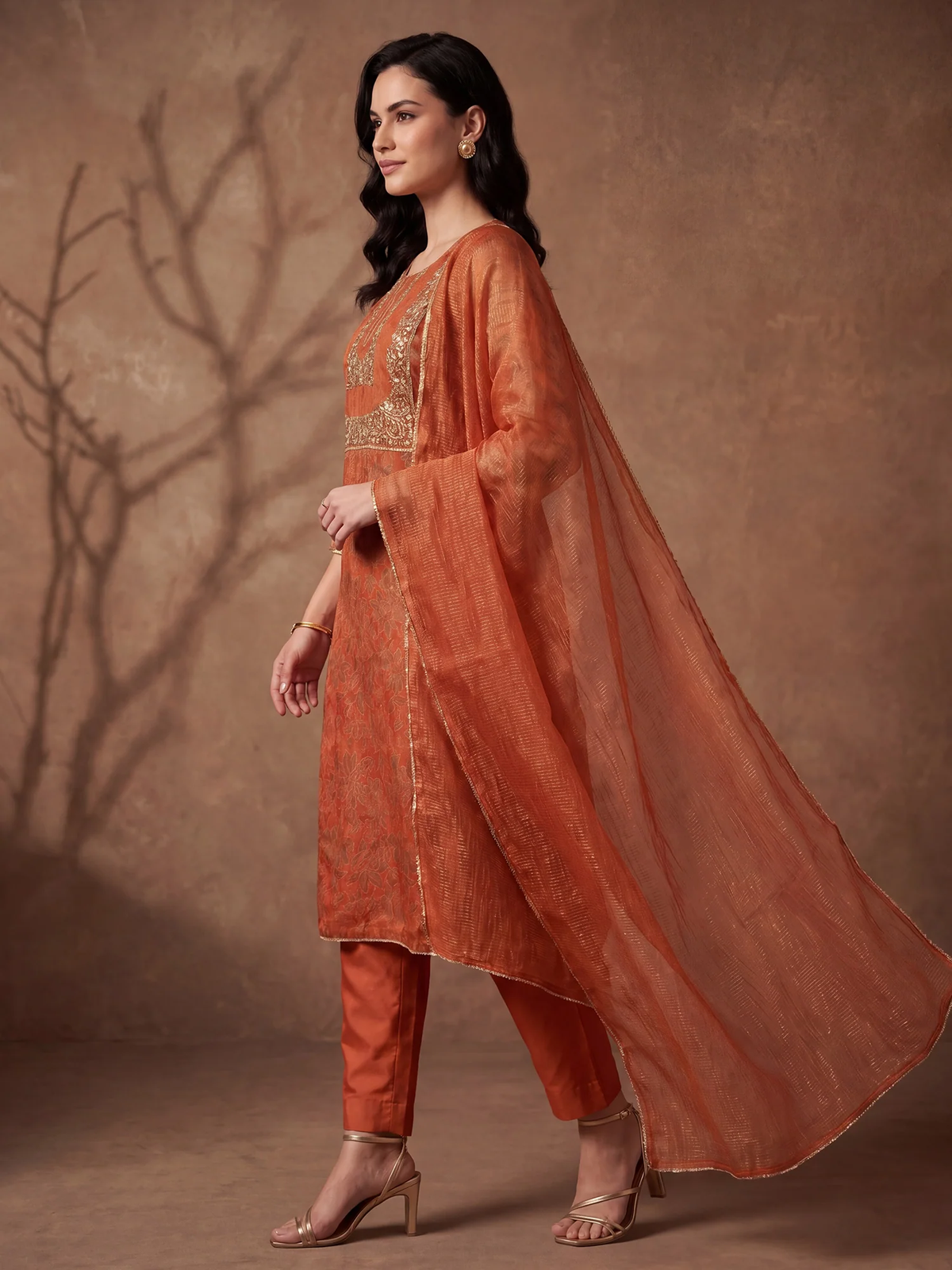 A woman models a traditional Indian salwar kameez outfit in rust orange, featuring an embroidered kameez kurta, matching trousers, and a sheer dupatta, set against a textured brown studio backdrop.