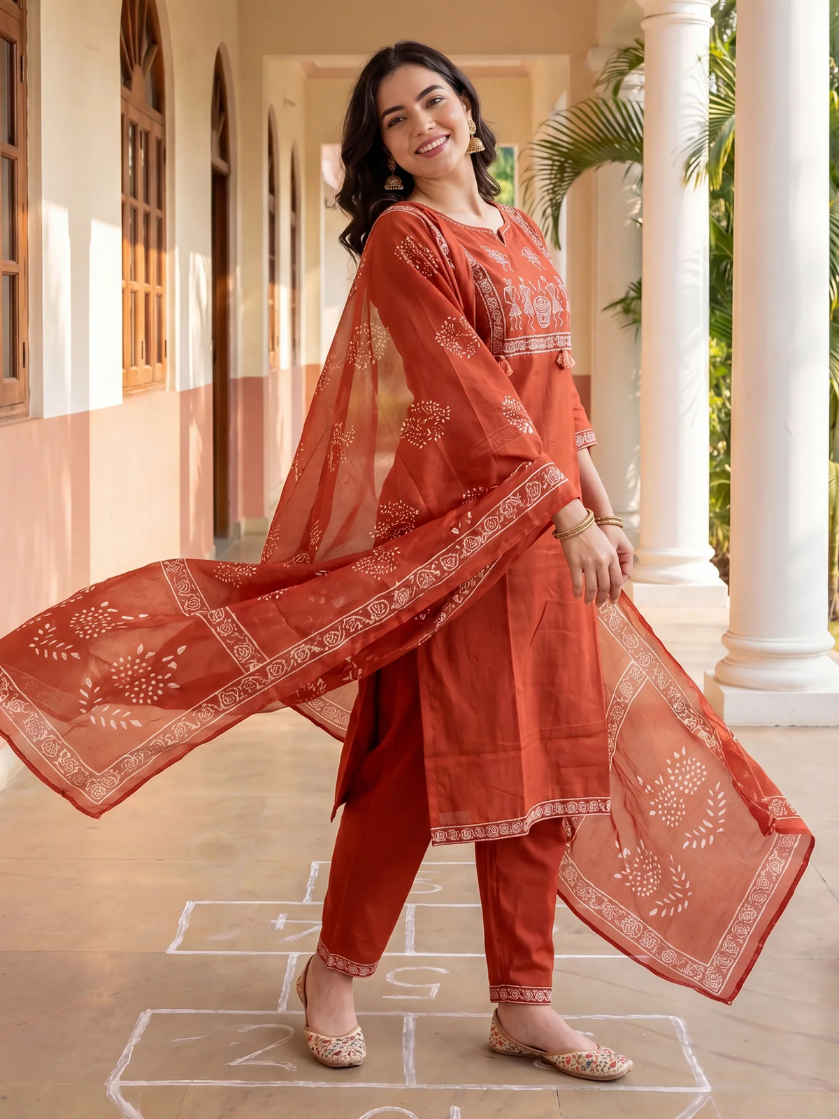 Woman in a traditional red outfit with white patterns standing in a sunlit room.