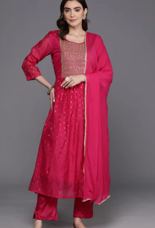 A woman modeling a bright pink traditional Indian outfit with a matching dupatta, featuring gold embroidery on the kurta and matching pink pants, set against a solid gray studio background.