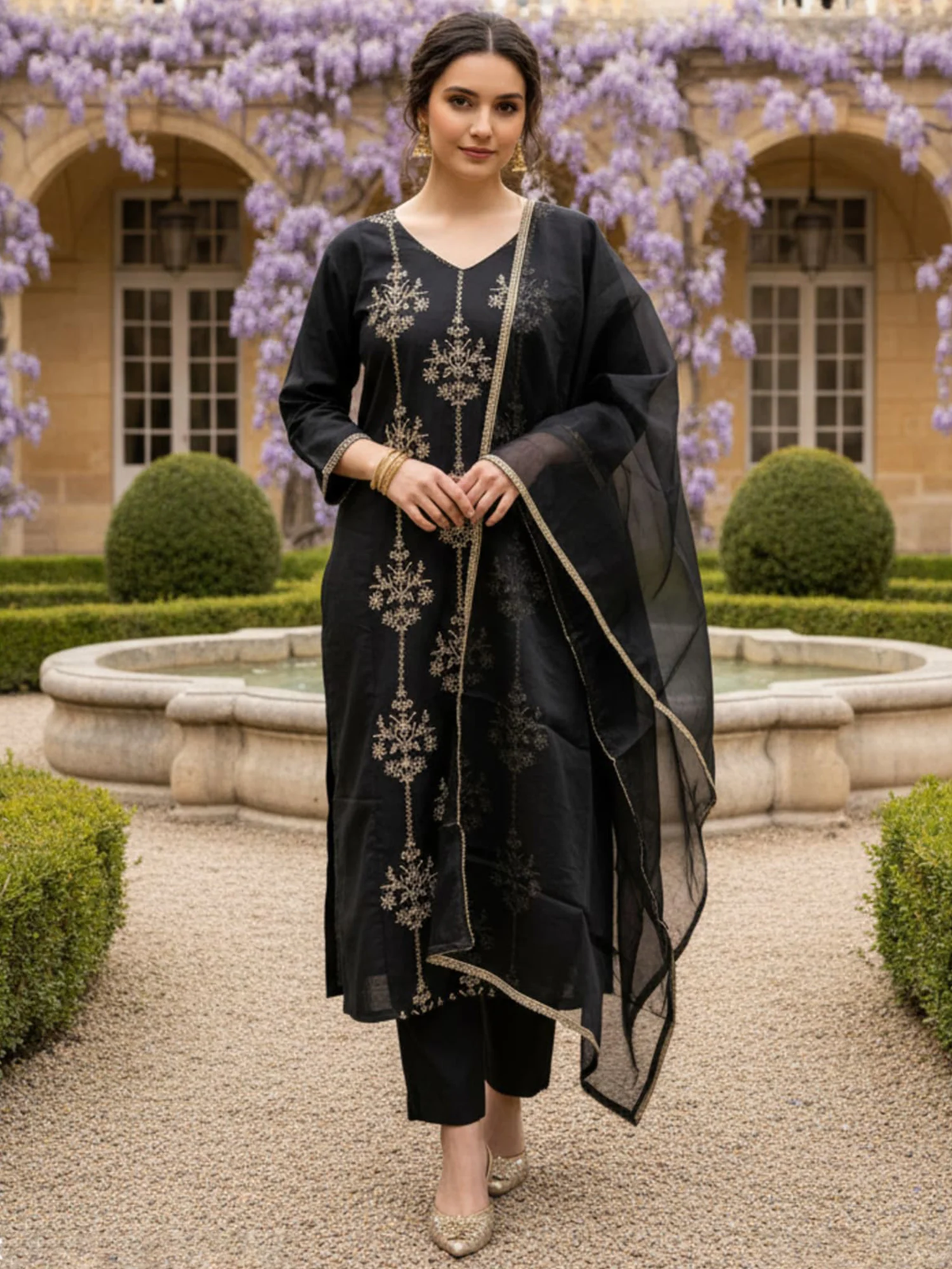 Woman in traditional black outfit with floral patterns standing in a garden with a fountain and purple flowers.