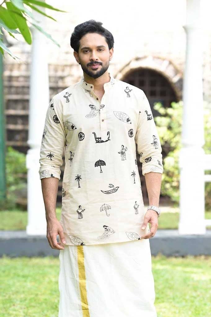 A man wearing a cream-colored kurta with black printed motifs and a white dhoti with a yellow stripe, standing outdoors in front of a white building with columns.