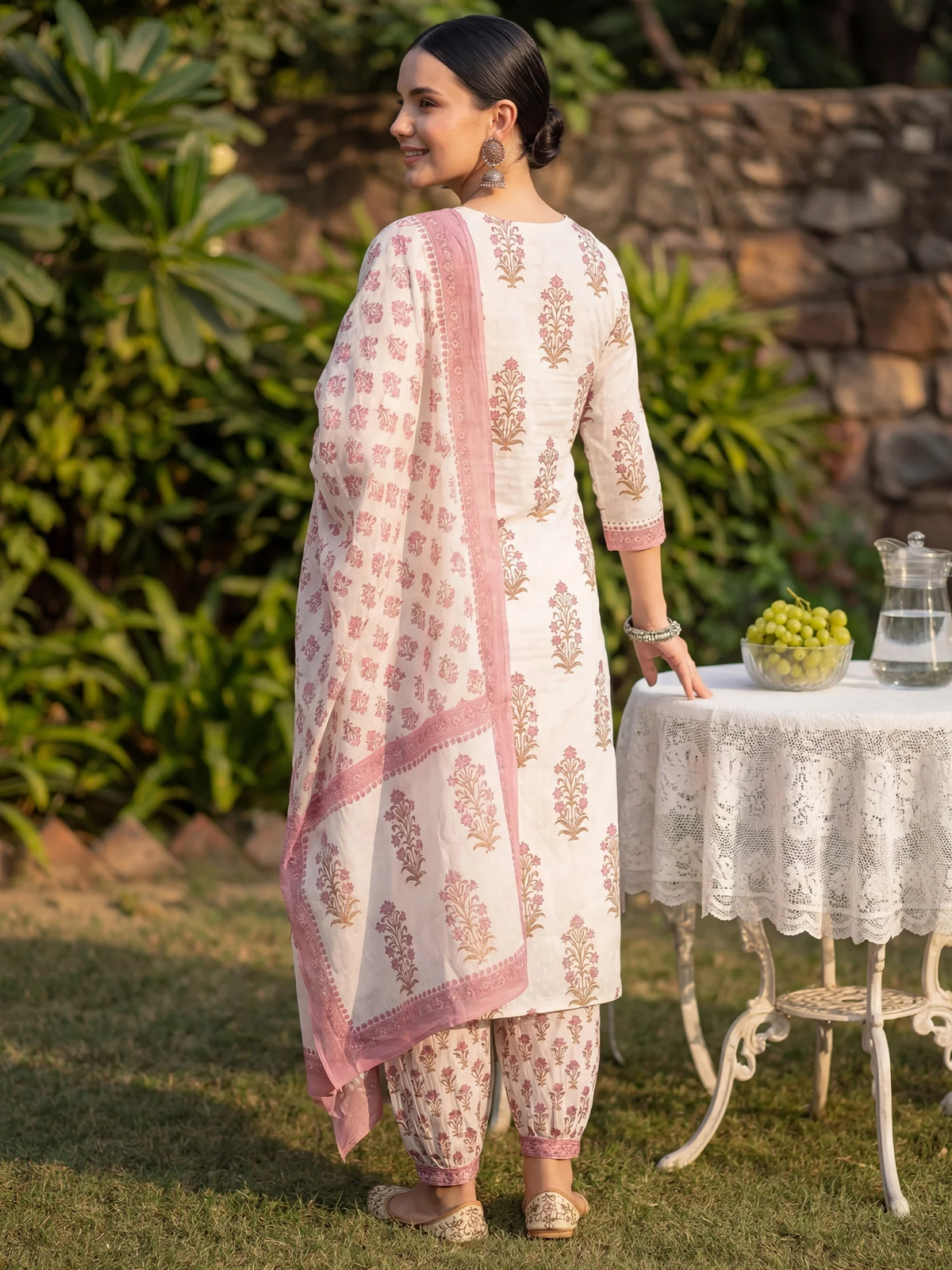 Woman in a traditional outfit standing outdoors near a table with a stone wall and greenery in the background.