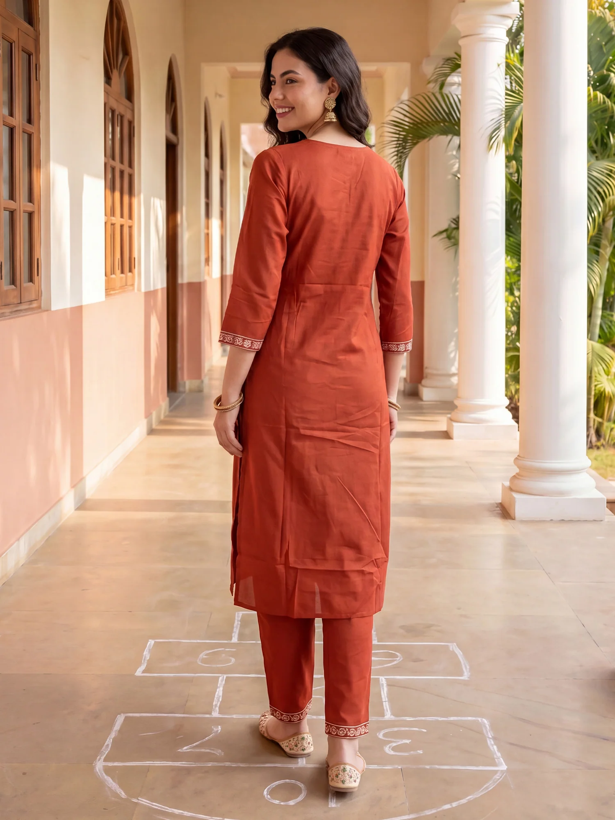 Woman in a rust-colored traditional outfit standing on a sunlit corridor with columns and greenery.