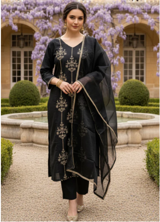 A woman stands in a garden courtyard wearing a black traditional outfit with beige embroidery and a sheer black dupatta with beige trim. The background features a stone building, trimmed hedges, and purple wisteria flowers.