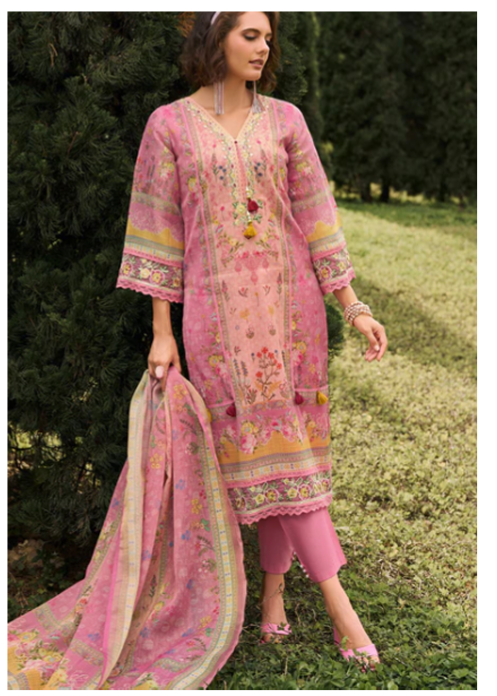 A woman stands outdoors on grass in front of dark green trees, wearing a pink embroidered traditional outfit with a matching dupatta. The outfit has floral and geometric patterns in pink, gold, and green, with pink pants and pink pointed shoes.