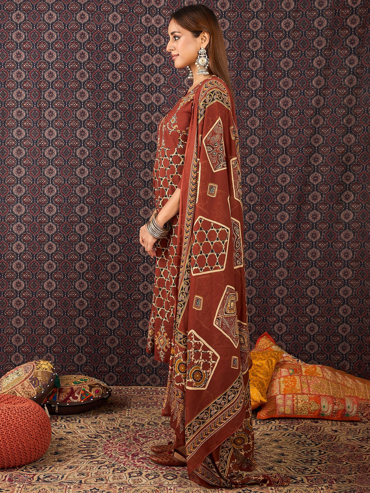 Woman in a patterned dress standing against a decorative wall with cushions on the floor.