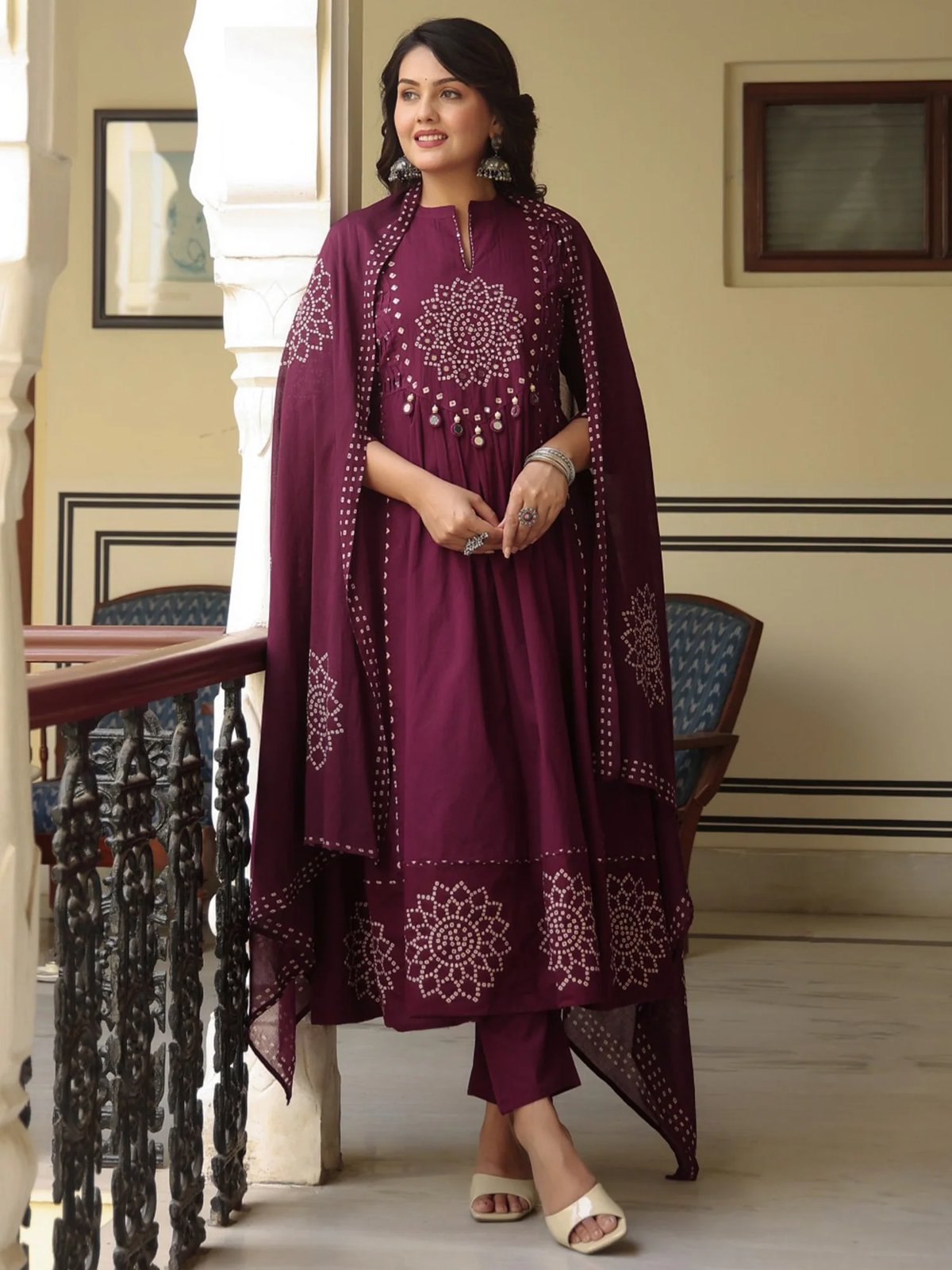 A woman models a deep maroon traditional Indian outfit with matching dupatta, featuring white floral embroidery and mirror work on the kurta and dupatta, set against an indoor architectural background.