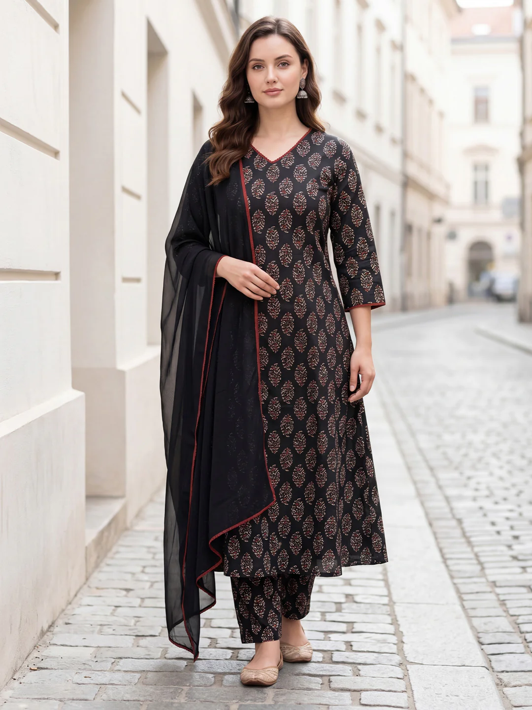 A woman stands on a cobblestone street wearing a black printed kurta set with a matching dupatta. The kurta has a repeating floral pattern in dark brown and beige, with red piping accents, and is paired with matching pants.