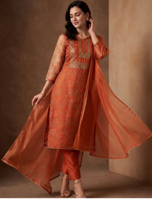 A woman models a traditional Indian salwar kameez suit in a warm rust-orange color with intricate gold embroidery on the chest, paired with matching pants and a sheer orange dupatta, set against a textured brown studio backdrop.