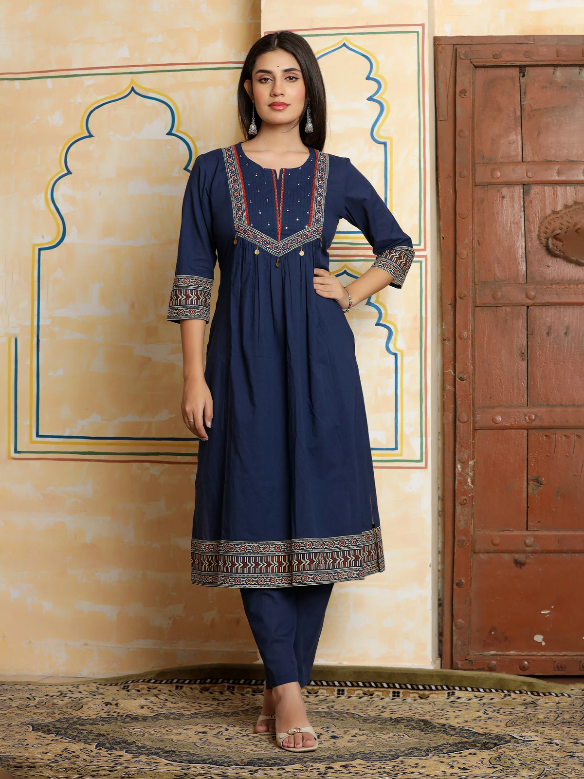 Woman wearing a navy blue traditional outfit with intricate patterns in front of a decorative wall.