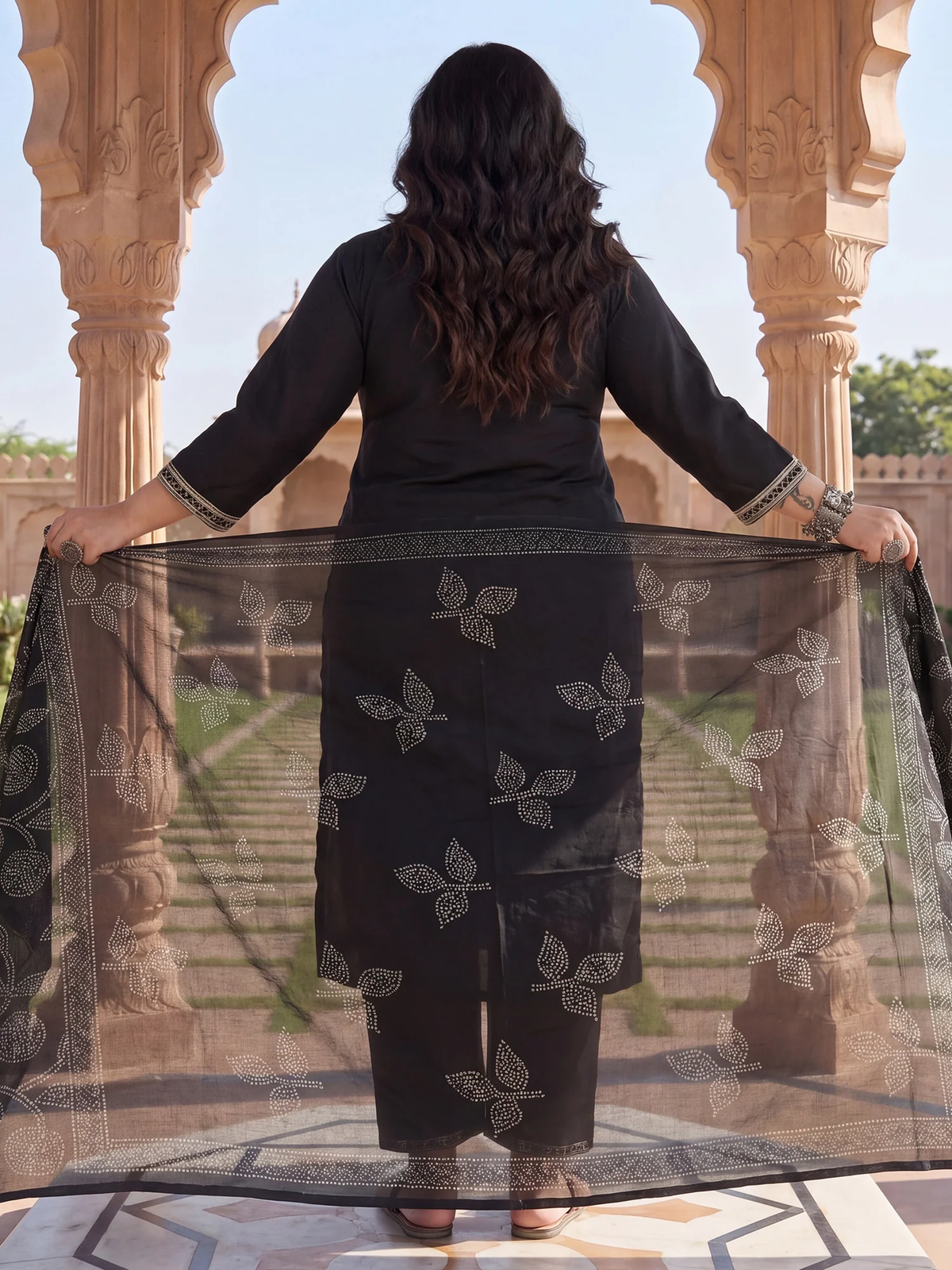 Woman in black traditional outfit holding a sheer fabric with intricate patterns in an outdoor setting.